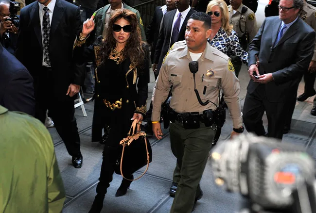 A woman in black attire, escorted by a security officer, walks through a crowd, surrounded by others.