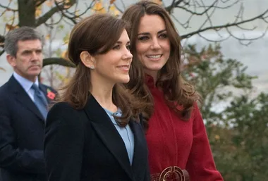 Two women walking outdoors and smiling, with a man in the background, trees and autumn leaves around.