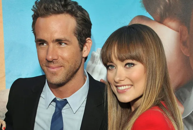 Couple smiling at a red carpet event, with a blue and beige background poster behind them.