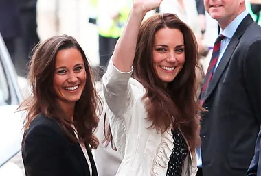 Two women smiling and waving at an outdoor event, surrounded by people.