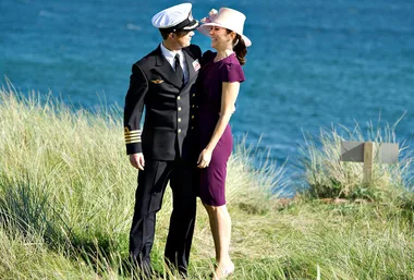 Air force officer in uniform and woman in purple dress embrace by sea, smiling at each other.