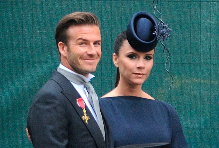 A man and woman in formal attire; the woman wears a navy dress and a small hat with adornments.