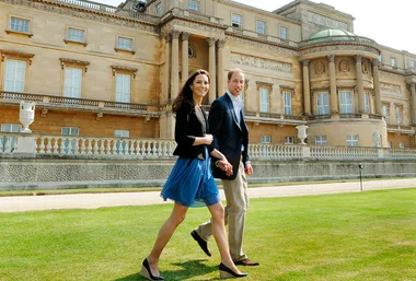 A couple holding hands, walking on the lawn in front of a large, elegant building.