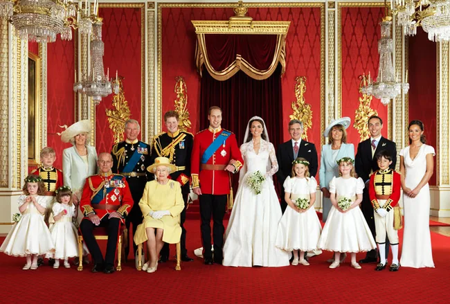 A royal family group portrait in formal attire in an ornate hall with red and gold decor.