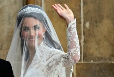 Kate Middleton waving in a lace wedding dress with veil and tiara against a stone backdrop.