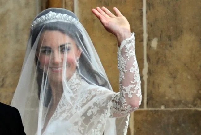 Kate Middleton waving in a lace wedding dress with veil and tiara against a stone backdrop.