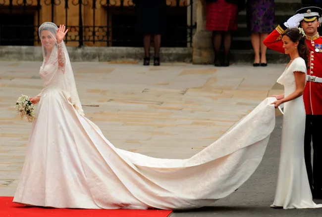 Bride in a long white dress waves, while bridesmaid holds her train on a red carpet outside a large building.