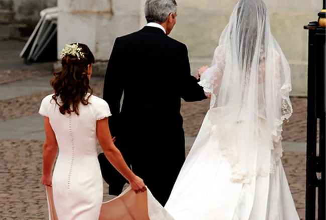 A bride in a white gown and veil walks with two people, one holding her train, on a cobblestone path.