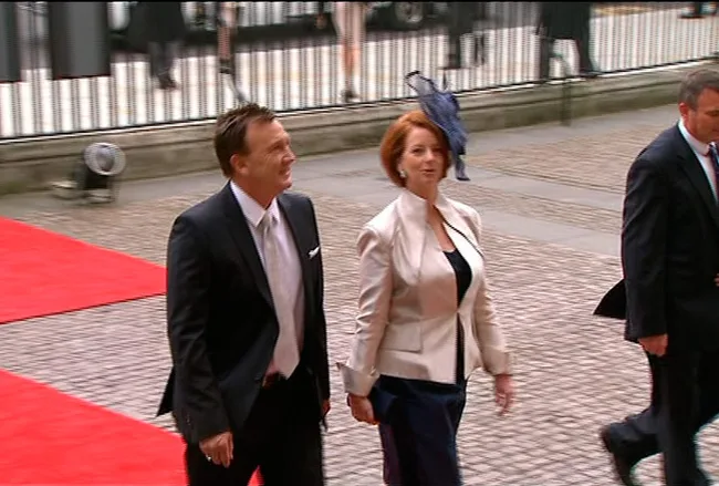 Two people walking on a red carpet outside, dressed in formal attire, with a blurred background of a fence and attendees.