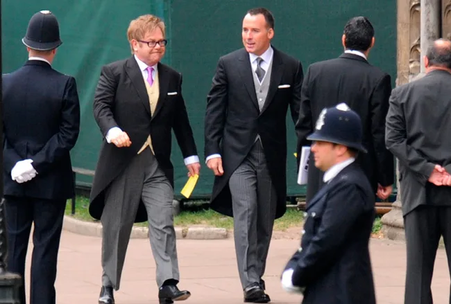 Two men in morning suits walking, holding paper. Police officers and other men in suits are visible in the background.