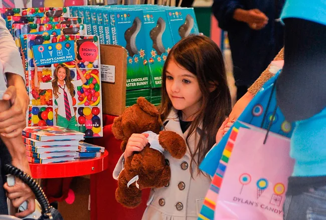 Young girl holding a teddy bear in a candy store with colorful boxes and bags around her.