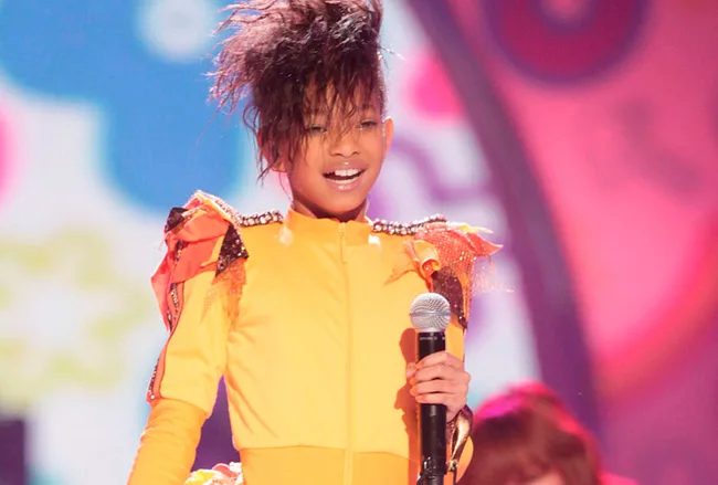 Singer in a vibrant yellow and orange outfit holding a microphone on stage with colorful background.