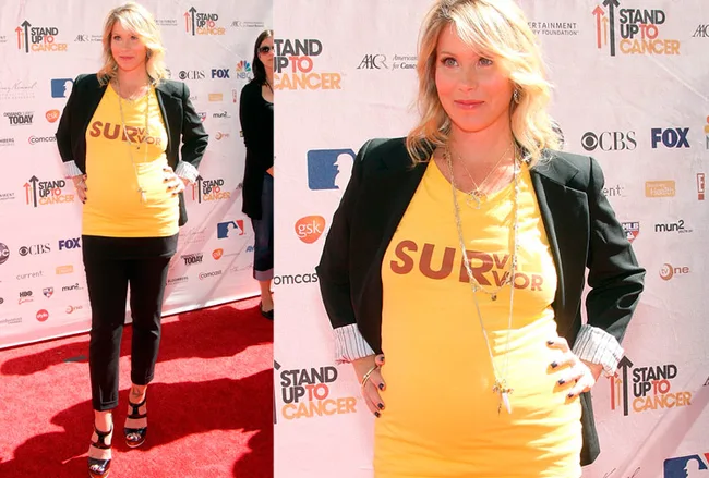 A pregnant woman in a yellow "Survivor" shirt and black blazer poses on a Stand Up To Cancer event red carpet.