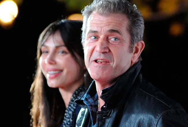 Two people standing together, one smiling and the other speaking, both in evening attire, with blurred lights in the background.
