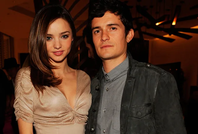 Two people posing together at an indoor event, both looking at the camera, with warm lighting in the background.