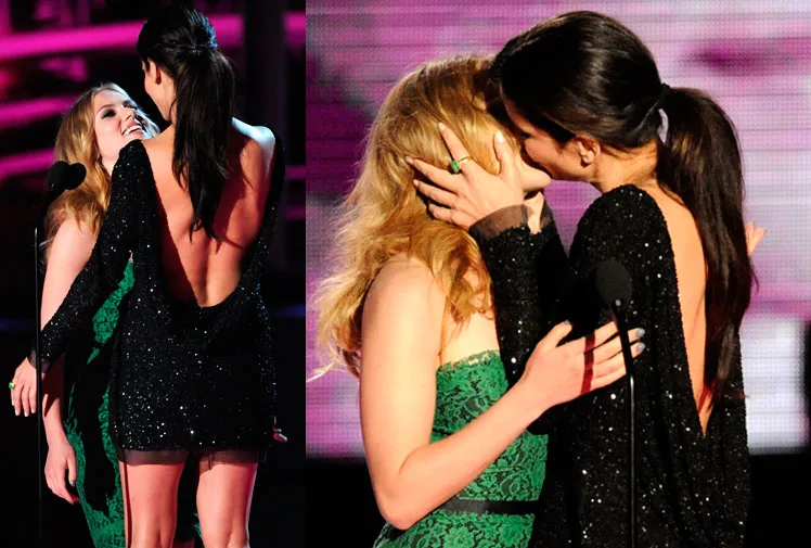 Two women in elegant dresses share a kiss on stage at an award event, with vibrant lighting in the background.