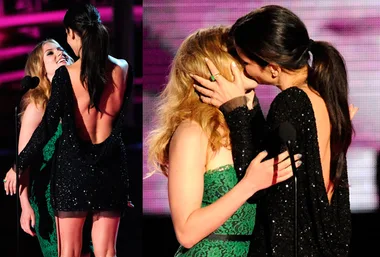 Two women in elegant dresses share a kiss on stage at an award event, with vibrant lighting in the background.