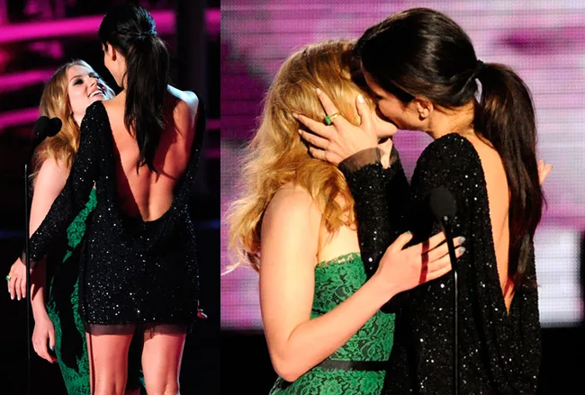 Two women in elegant dresses share a kiss on stage at an award event, with vibrant lighting in the background.