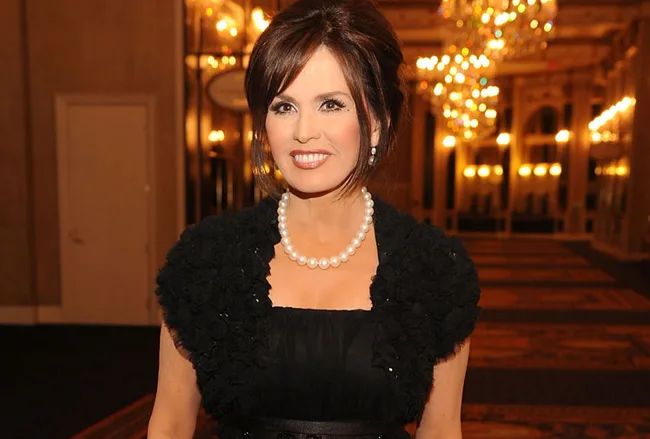 A woman in a black dress and pearl necklace smiling with chandeliers in the background.