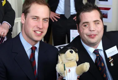 Prince William with a man holding a stuffed dog wearing sunglasses and a medal at a public event.