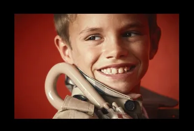 A young boy smiles widely, wearing a beige jacket with a coat hanger visible, against a red background.