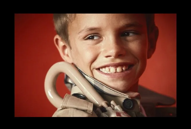 A young boy smiles widely, wearing a beige jacket with a coat hanger visible, against a red background.