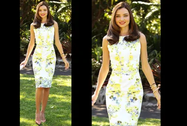 A woman in a floral dress stands outdoors in a garden, sunlight highlights her.