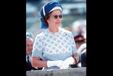 Woman in a blue hat and white gloves, wearing sunglasses and a patterned dress, standing outdoors.