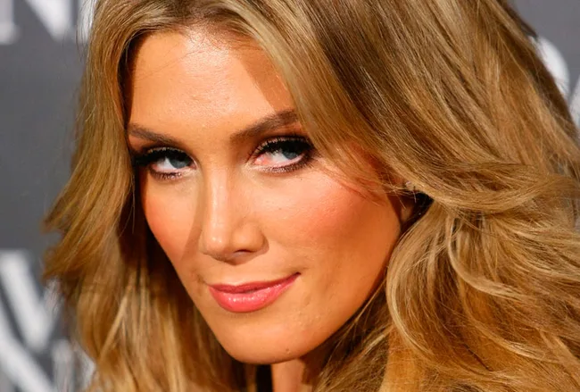 A person with long, wavy blonde hair and defined eyelashes, wearing subtle makeup, looking confidently at the camera.