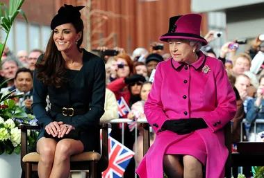 Royal figures sitting together at a public event, surrounded by crowd with Union Jack flags in background.