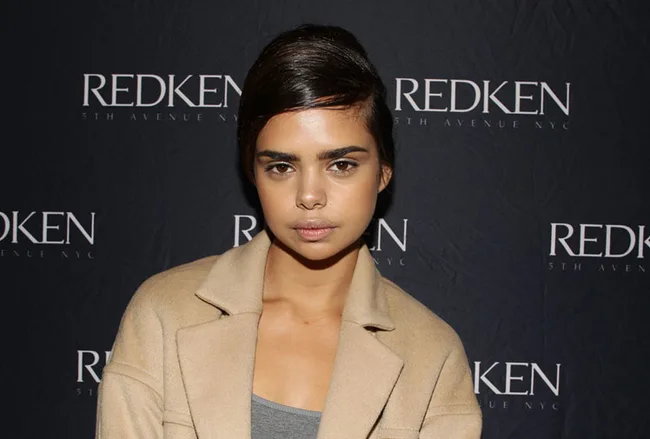 Person with sleek hairstyle and beige coat at a Redken event backdrop.