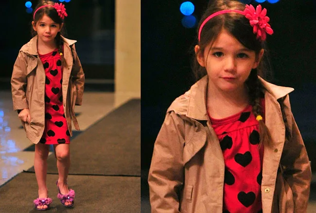 Young girl in a red heart-patterned dress and beige coat, with a red flower headband, walking indoors.