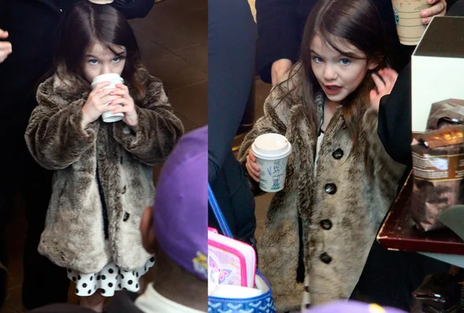 Girl in a faux fur coat holding a drink in a coffee shop, with people around her.