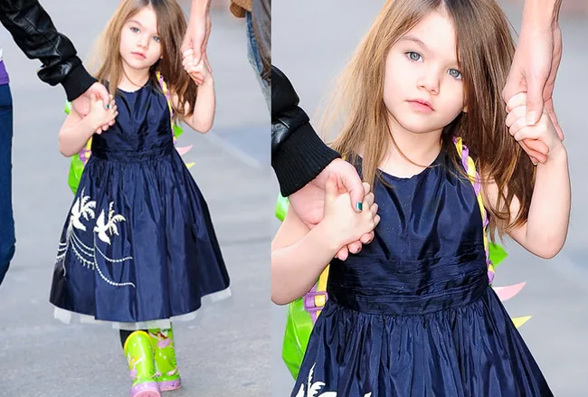 Young girl in a blue dress with a floral pattern, holding an adult's hand, wearing green boots and a backpack.