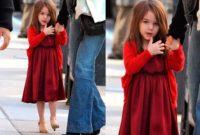 A young girl in a red dress and cardigan holding an adult's hand, walking on a sidewalk.