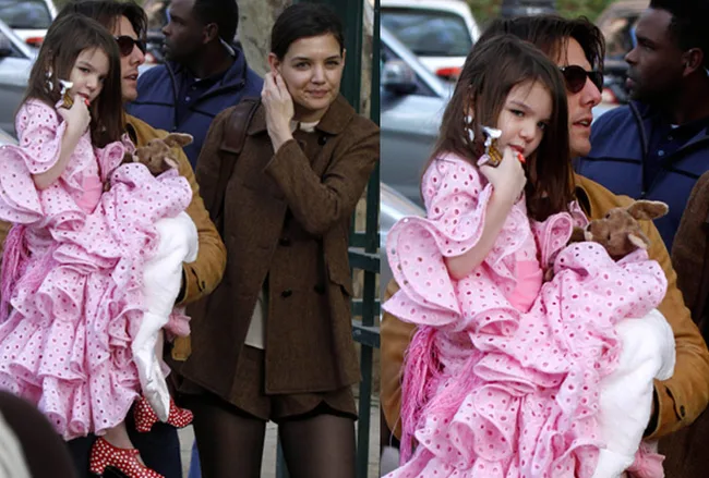 A family walks outside, a child in a pink dress held by an adult, another adult beside them.