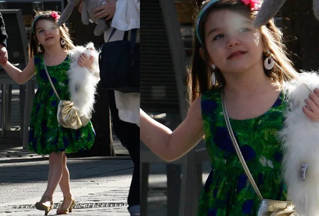 Young girl in a green dress and gold shoes, holding a fluffy toy and a gold purse, walking outdoors.