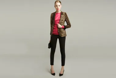 Model in leopard print blazer, pink top, black pants, and heels holding a clutch against a gray background.