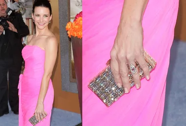 A woman in a bright pink dress holding a sparkly clutch, posing at an event with photographers in the background.