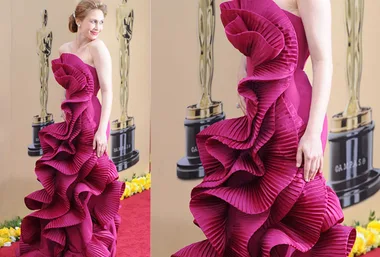 A person in a vibrant pink ruffle gown on a red carpet, with an Oscar statue in the background.