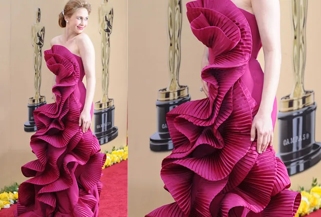 A person in a vibrant pink ruffle gown on a red carpet, with an Oscar statue in the background.