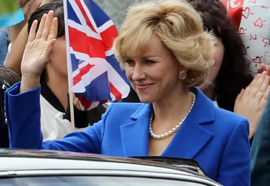 Actress waves in blue suit with pearls, holding a UK flag, portraying royalty.