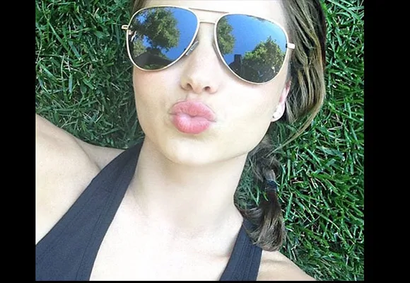 Selfie of a person lying on grass, wearing sunglasses and a dark tank top, making a kissy face.