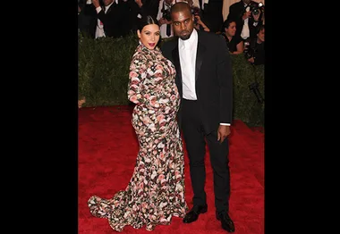 A couple poses on a red carpet; the woman wears a floral dress, and the man is in a black tuxedo.