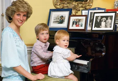 Princess Diana smiles as she stands by two young boys playing the piano, with framed photos on top.