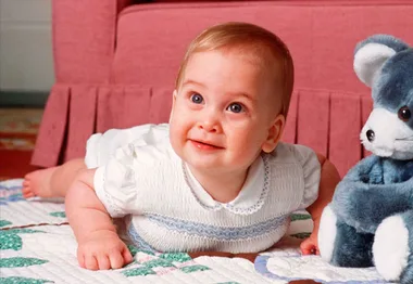 Baby lying on a quilted blanket, smiling, with a blue teddy bear nearby.