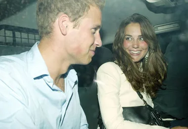 Two people smiling at each other while sitting in a car, seen through a window.
