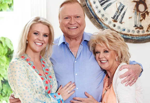 Three smiling people embracing indoors, standing in front of a large wall clock.