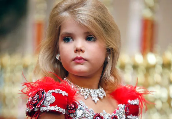 A young girl in a sparkling red dress with blond hair and jewelry, looking away from the camera.