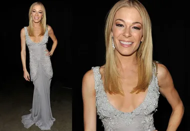 Blonde woman in a silver gown with intricate details, smiling and posing in a dimly lit setting.